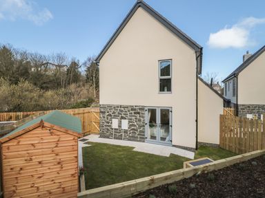 A backyard with a grass lawn a wooden shed a paved area and a two-story house with stone and beige walls at High Tide House in Craster