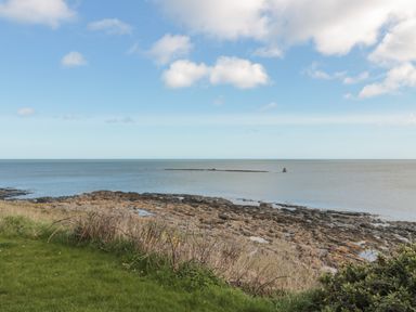 A rocky shoreline with grass and shrubs near the ocean under a partly cloudy sky at High Tide House in Craster