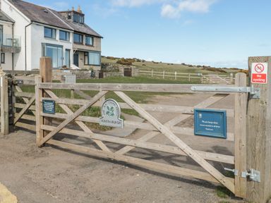A wooden gate with multiple signs including no cycling and National Trust Dunstanburgh in front of houses and a grassy field at High Tide House in Craster