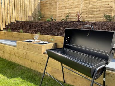 A black barbecue grill in a garden with wooden seating and a table set with plates and glasses at High Tide House in Craster