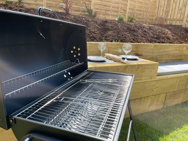 An outdoor barbecue grill with open lid next to a wooden bench with cushions and a table set with plates wine glasses and bowls at High Tide House in Craster