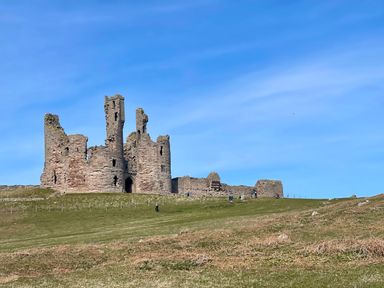 A ruined stone castle on a grassy hill under a blue sky at High Tide House in Craster