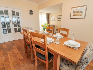 A dining room with a wooden table set with plates glasses and a vase of flowers with wooden chairs and patterned armchairs at Beach Cove View in Newbiggin-by-the-Sea