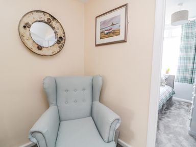 A corner with a light blue armchair a round wall mirror and a framed picture with a glimpse into a bedroom with plaid curtains and a gray carpet at Beach Cove View in Newbiggin-by-the-Sea