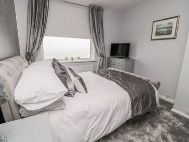 A bedroom with a bed next to a window with curtains and a TV on a chest of drawers at Beach Cove View in Newbiggin-by-the-Sea