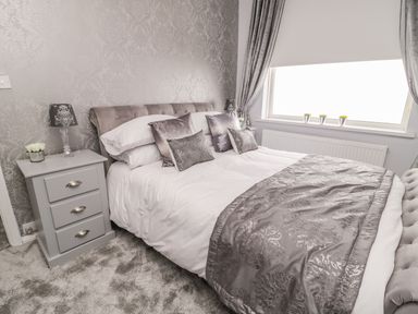 A bedroom with a bed with grey and white bedding a grey nightstand with a lamp and flowers and a window with curtains at Beach Cove View in Newbiggin-by-the-Sea