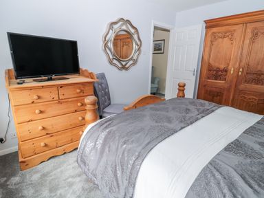 A bedroom with a wooden dresser holding a television a gray chair a wooden wardrobe a bed with patterned bedding and a wall mirror at Beach Cove View in Newbiggin-by-the-Sea