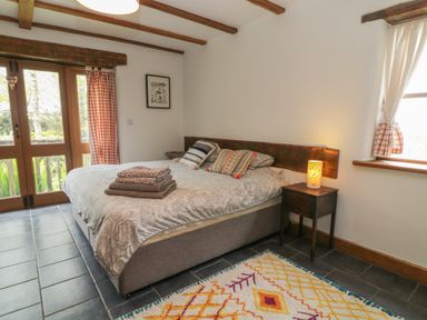 A bedroom with a bed with pillows and folded towels a bedside table with a lamp and windows with curtains at Woodcutters in Rezare near Launceston