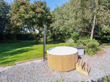 A wooden hot tub on gravel with wooden steps surrounded by grass trees and a bench in a garden at Woodcutters in Rezare near Launceston