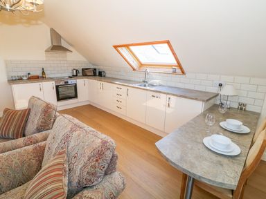 A kitchen with white cabinets a countertop window and a small dining area with set tableware at Llain Haf in Llangefni
