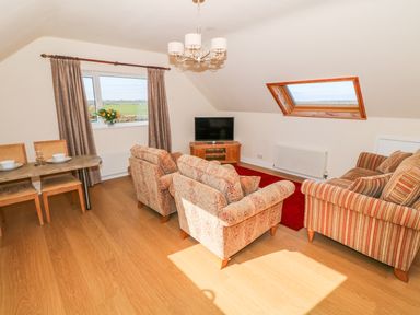 A living room with a dining table with chairs two patterned armchairs a striped sofa a television on a wooden stand and windows with curtains at Llain Haf in Llangefni