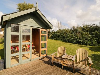 A wooden garden shed with open doors and two wooden chairs on a deck with grass and bushes in the background at Llain Haf in Llangefni