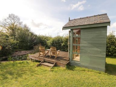 A small green wooden cabin with glass doors and two wooden chairs on a deck in a garden at Llain Haf in Llangefni
