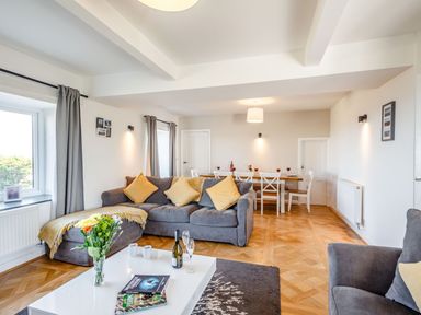 A living room with grey sofas yellow cushions a white coffee table with flowers and books and a dining table with white chairs at Tyn Y Berth in Benllech