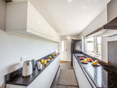 A narrow kitchen with white cabinets black countertops a kettle various fruits and drinks near a window and a black refrigerator at Tyn Y Berth in Benllech