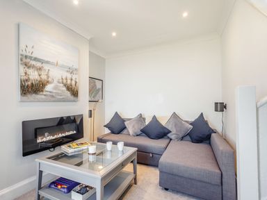 A living room with a gray sectional sofa with cushions a glass coffee table with books and cups a wall-mounted electric fireplace and a painting above at Tyn Y Berth in Benllech