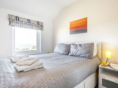 A bedroom with a patterned bedspread and pillows towels on the bed a nightstand with a lamp a glass of juice and an open book sunset artwork above the bed and a window with a view at Tyn Y Berth in Benllech
