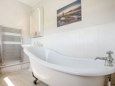 A bathroom with a white freestanding bathtub with claw feet a wall-mounted heated towel rack a pedestal sink and a scenic coastal photograph on the wall at Tyn Y Berth in Benllech
