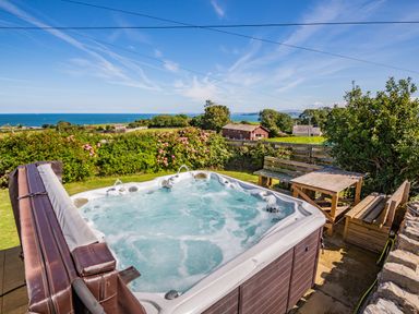 An outdoor hot tub with wooden benches and a table overlooking a garden and sea at Tyn Y Berth in Benllech