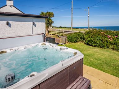 An outdoor hot tub on a patio next to a white building with a garden and ocean view at Tyn Y Berth in Benllech