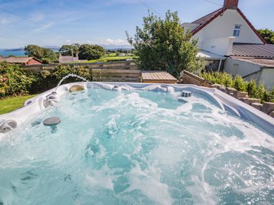 An outdoor hot tub with bubbling water and a wooden fence with bushes and a house in the background at Tyn Y Berth in Benllech