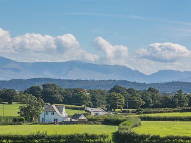 A rural landscape with a white house surrounded by green fields and trees with mountains in the background at Tyn Y Berth in Benllech