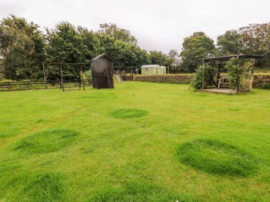 A garden with a swing set a small dark wooden playhouse a play structure with a slide and a wooden gazebo with plants at Shepherds Hut - The Hurdle in Rosemarket