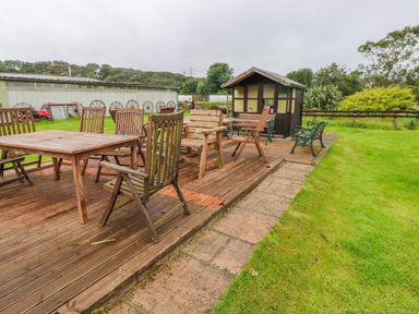 An outdoor wooden deck with wooden chairs and tables a small shed and green benches on grass at Shepherds Hut - The Hurdle Rosemarket