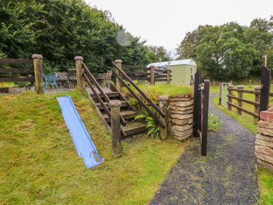 An outdoor seating area with tables and chairs behind wooden stairs and a small blue slide in a grassy yard at Shepherds Hut - The Hurdle in Rosemarket