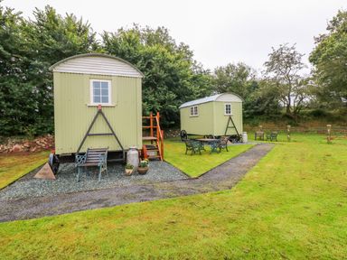 Two green shepherd huts with wooden stairs and benches on a grassy area with trees in the background at Shepherds Hut - The Hurdle in Rosemarket