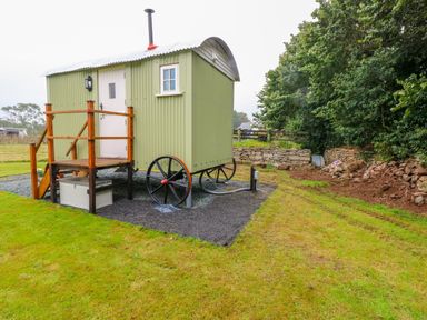 A green shepherds hut with wheels on a gravel area beside grass and trees at Shepherds Hut - The Hurdle in Rosemarket