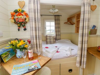 A small bedroom with a bed towels and curtains with a table holding flowers wine glasses and a magazine at Shepherds Hut - The Hurdle in Rosemarket
