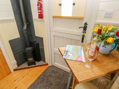 A corner with a small stove beside a wooden table with wine glasses a bottle magazine and flowers at Shepherds Hut - The Hurdle in Rosemarket