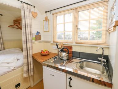 A small kitchen area with a sink stove kettle and fruit bowl next to a bed separated by a curtain at Shepherds Hut - The Hurdle in Rosemarket