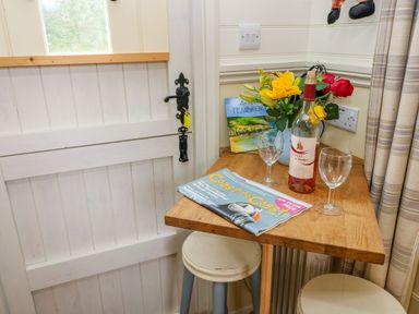 A small wooden table with a wine bottle flowers two glasses and a magazine next to a white door with stools at Shepherds Hut - The Hurdle in Rosemarket