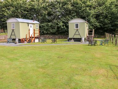 Two small huts with steps on a grass lawn with benches and tables surrounded by trees at Shepherds Hut - The Hurdle in Rosemarket