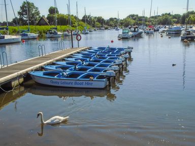 A row of blue boats tied to a dock with a swan swimming nearby and sailboats in the background at 32 Cranleigh Close in Tuckton