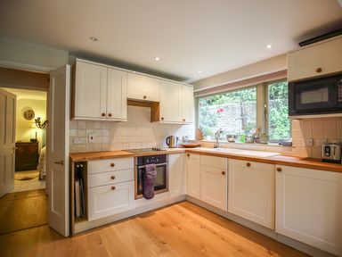 A kitchen with white cabinets a wooden countertop an oven a microwave and a window overlooking a garden at All Souls Cottage in Eastleach Cotswolds