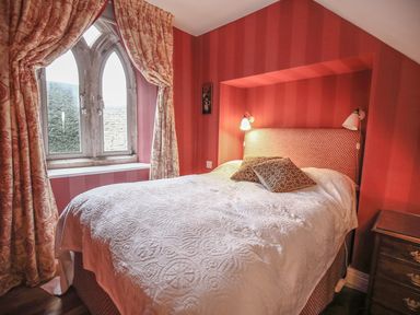 A bedroom with a patterned bedspread and pillows near a stone arched window with curtains at All Souls Cottage in Eastleach Cotswolds