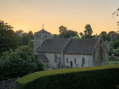 A stone church with a small tower and arched windows surrounded by trees at sunset at All Souls Cottage in Eastleach Cotswolds