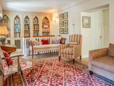 A sitting room with patterned armchairs a sofa built in bookcases and a red patterned rug at All Souls Cottage in Eastleach Cotswolds