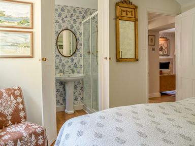 A bedroom with a bed and chair showing a bathroom with a sink and shower and a living room in the background at All Souls Cottage in Eastleach Cotswolds