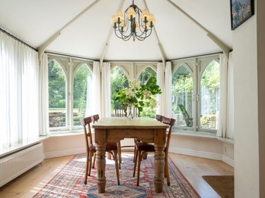 A dining area with a wooden table and four chairs under a chandelier in a room with arched windows and white curtains at All Souls Cottage in Eastleach Cotswolds