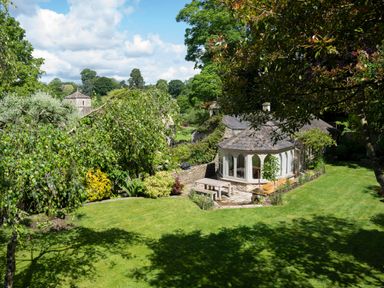 A garden with green lawn trees and a stone building with large windows at All Souls Cottage in Eastleach Cotswolds