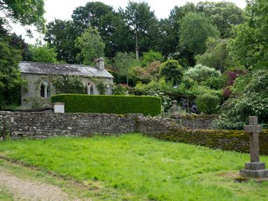 A stone cottage surrounded by trees and shrubs with a stone wall and graveyard in the foreground at All Souls Cottage in Eastleach Cotswolds