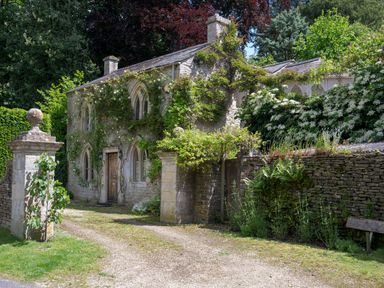 A stone cottage with climbing plants and bushes around a driveway at All Souls Cottage in Eastleach Cotswolds