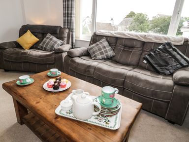 A living room with a wooden coffee table holding a tea set and cupcakes and two brown sofas with cushions at Ty Gilbert in Benllech