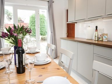 A dining area with a wooden table set with white plates bowls wine glasses and a bottle of wine next to a kitchen with grey cabinets and a countertop at Ty Gilbert in Benllech
