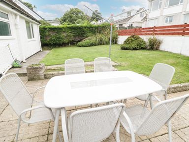 A patio area with a white metal table and six chairs next to a garden with grass and shrubs at Ty Gilbert in Benllech