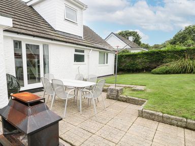 An outdoor patio with a metal table and six chairs next to a white house and a grassy garden at Ty Gilbert in Benllech